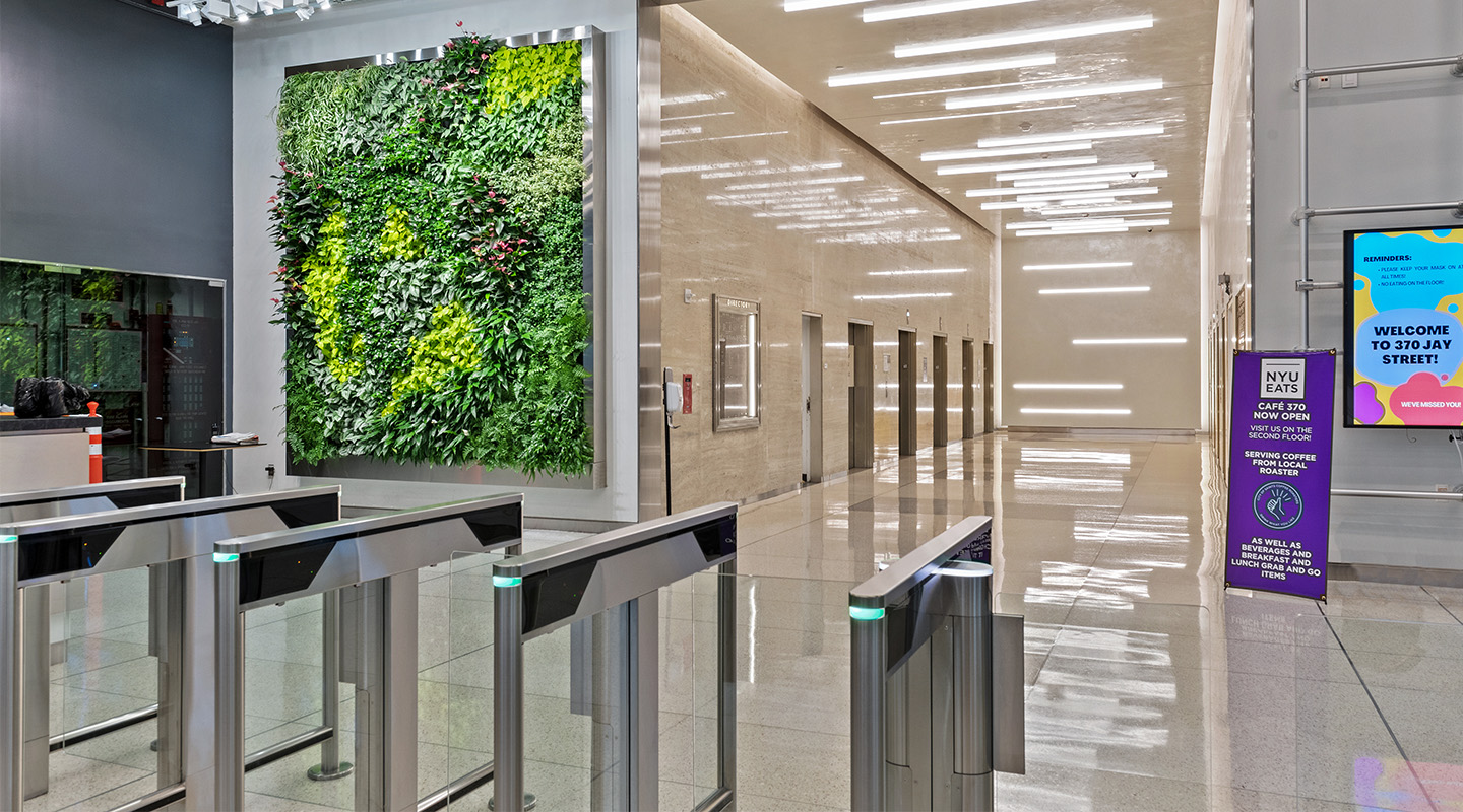 A modern building lobby with a large vertical living plant wall, shiny floors, security turnstiles, elevators at the end, and digital signs with information and welcome messages.