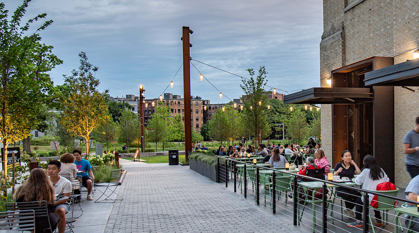 People dine outdoors at tables beside a modern building, with string lights overhead and trees and greenery in the background. The sky is cloudy, and the atmosphere is lively and relaxed.