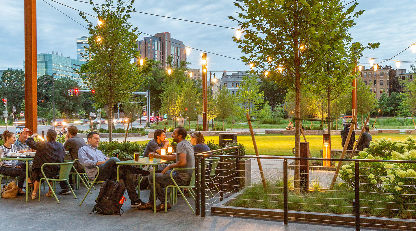 People sit at outdoor tables on a patio in an urban park, talking and eating under string lights during the evening. Green lawns, trees, and city buildings are visible in the background.