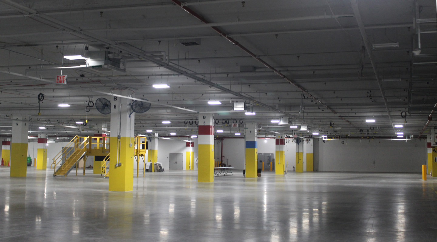 A large, empty indoor parking garage or warehouse with yellow pillars, concrete floors, overhead lighting, and a stairway leading to an upper platform. No vehicles or people are present.