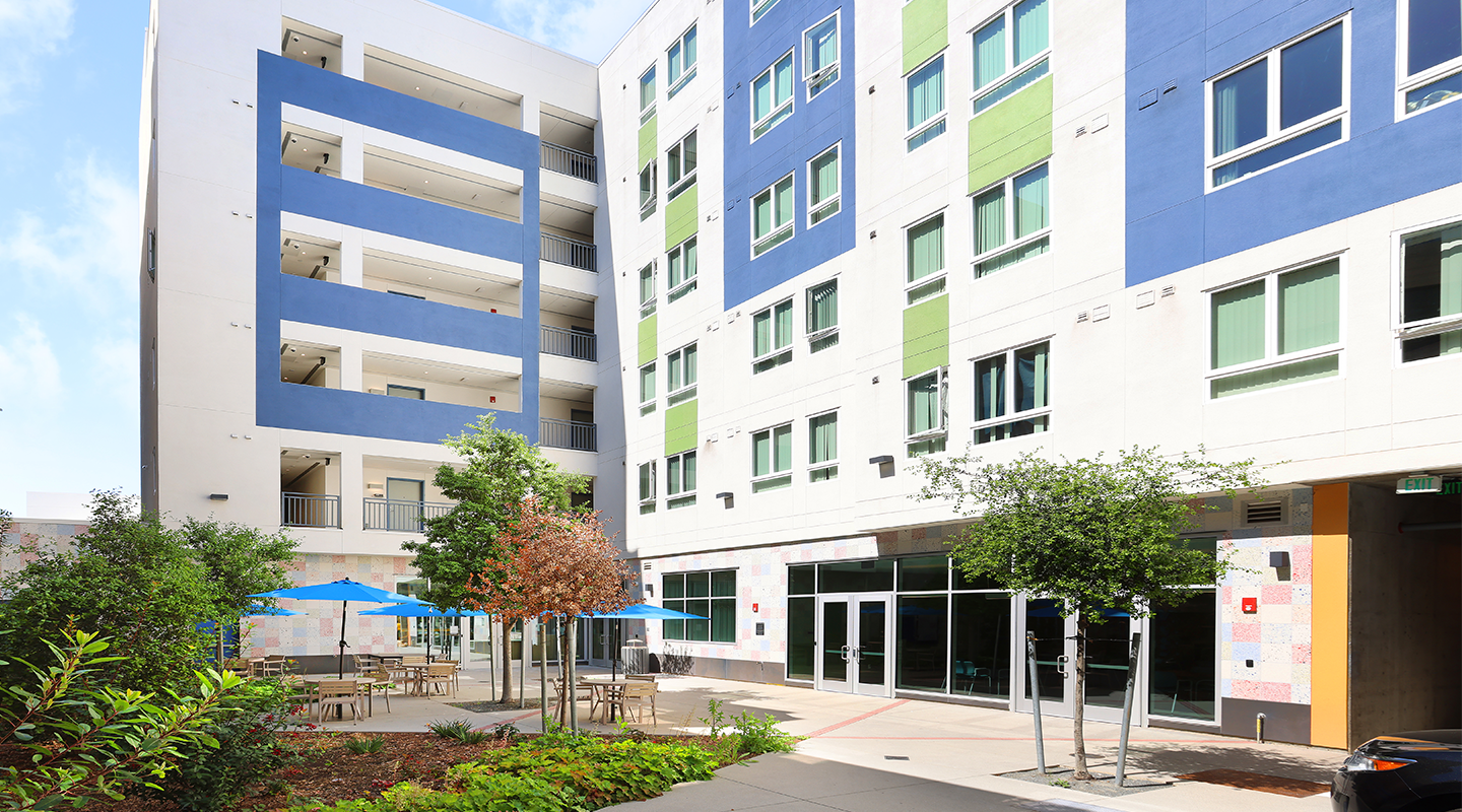 A modern apartment building with blue, green, and white panels, large windows, and a courtyard featuring outdoor tables, blue umbrellas, and trees. The setting is bright and inviting with clear weather.