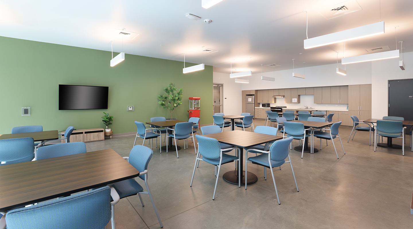 A modern break room with several tables and blue chairs, a green accent wall with a mounted TV, a popcorn machine, potted plant, kitchen area with cabinets, and bright ceiling lights.