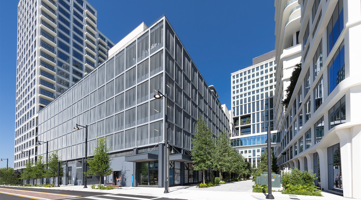 Street view of modern high-rise buildings with glass facades, trees lining the sidewalks, lamp posts, and a clear blue sky above.