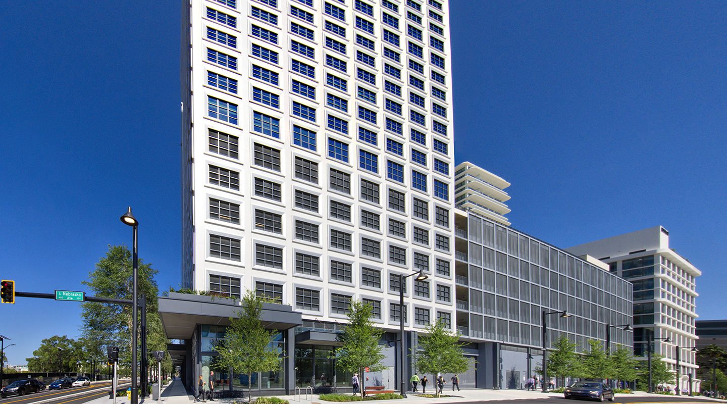 A modern high-rise building with a grid-like window pattern stands on a sunny street corner, surrounded by trees, pedestrians, and clear blue sky.
