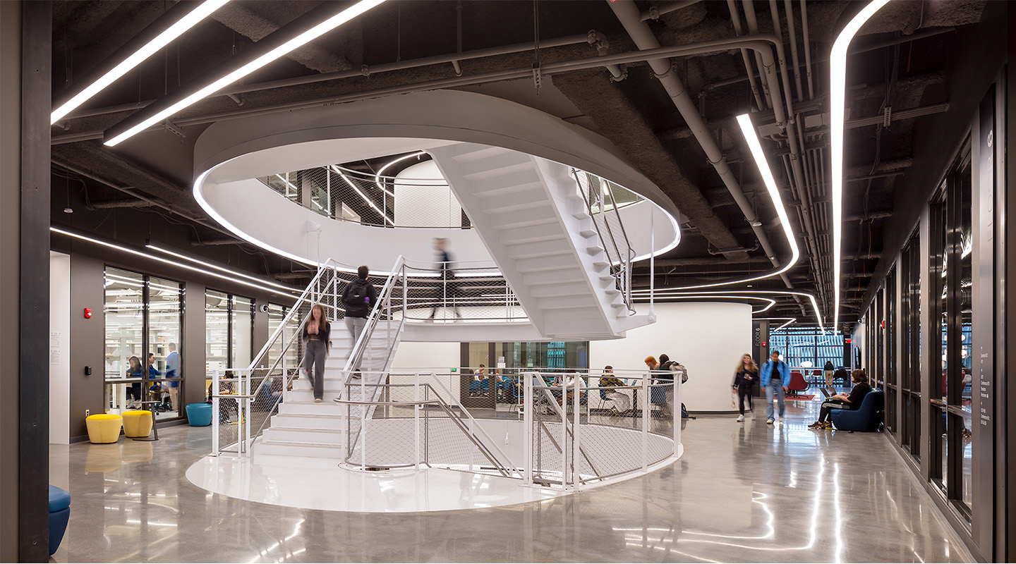 A modern office interior with a large circular white staircase in the center, people walking up and down the stairs, polished floors, glass walls, and LED strip lighting on the ceiling.