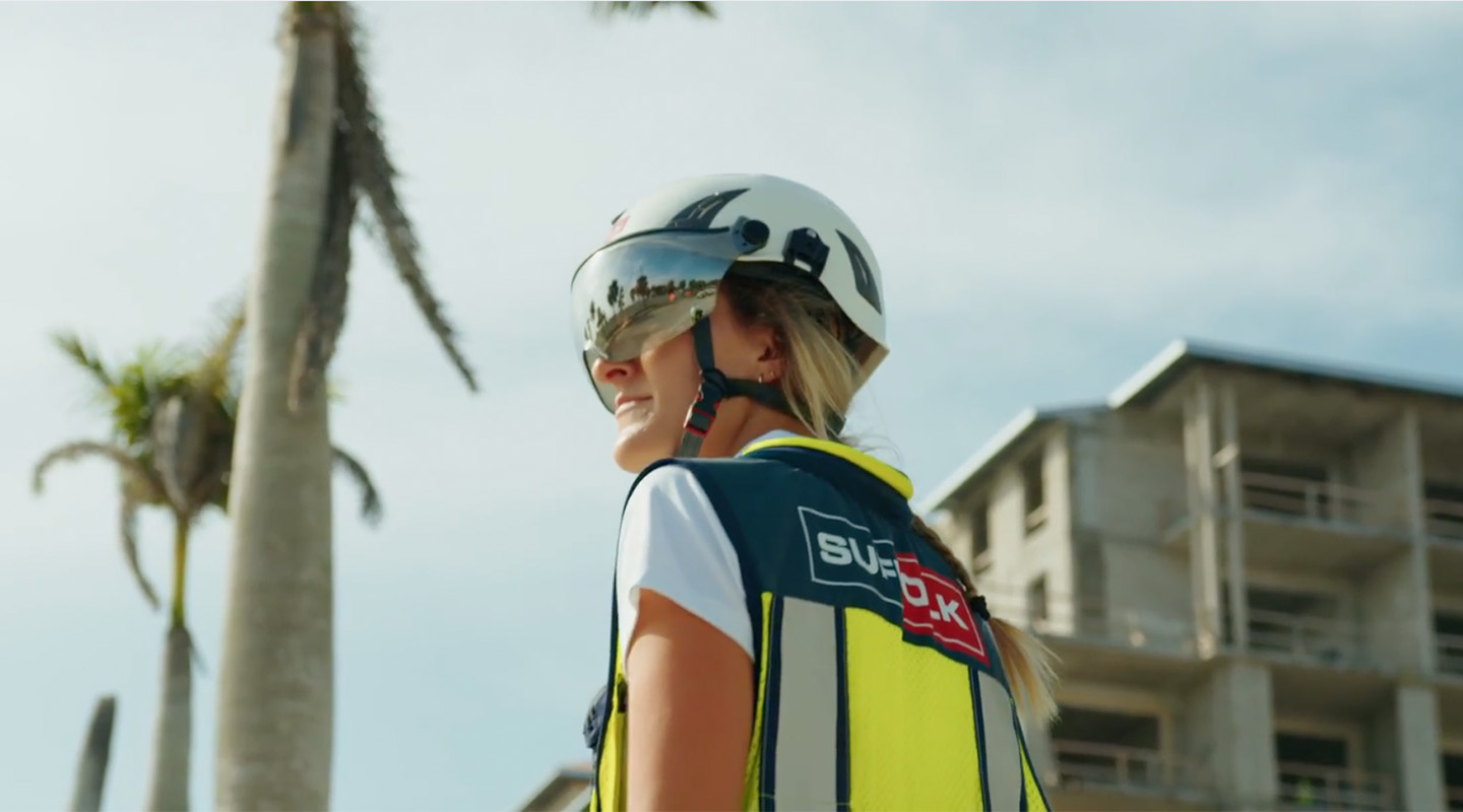 A woman in a hard hat, mirrored safety glasses, and a high-visibility vest stands outdoors near palm trees and an unfinished building, looking over her shoulder and smiling.