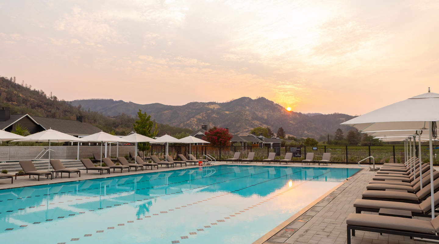A serene outdoor swimming pool lined with lounge chairs and umbrellas sits under a pastel sunset, with mountains and trees in the background. The calm water reflects the sky and surrounding landscape.