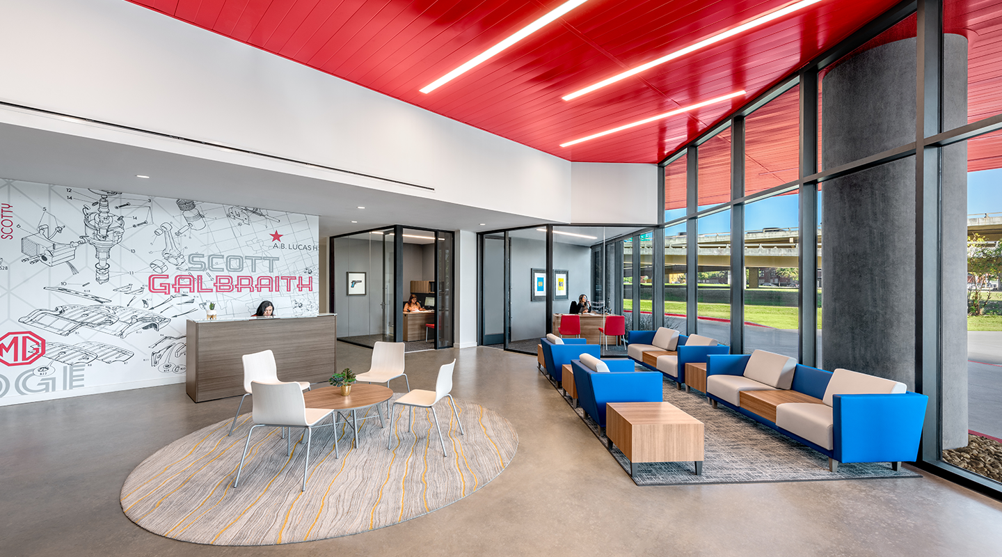 Modern office lobby with floor-to-ceiling windows, blue and beige seating, a circular rug with a table and chairs, a reception desk, and a wall with colorful graphics and text. Red ceiling panels add a vibrant touch.