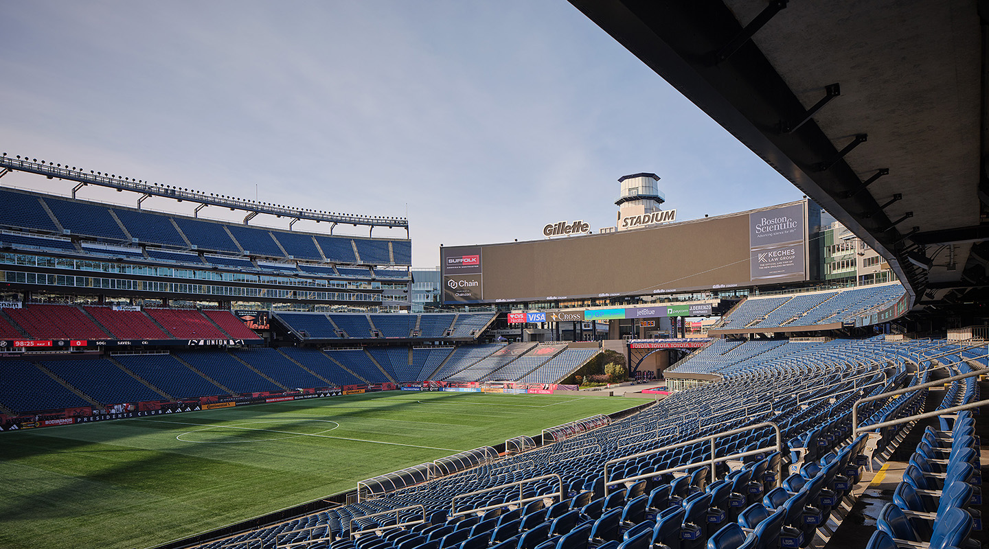 View of an empty Gillette Stadium with rows of blue seats, a large scoreboard, and a well-maintained green soccer field under a partly cloudy sky.