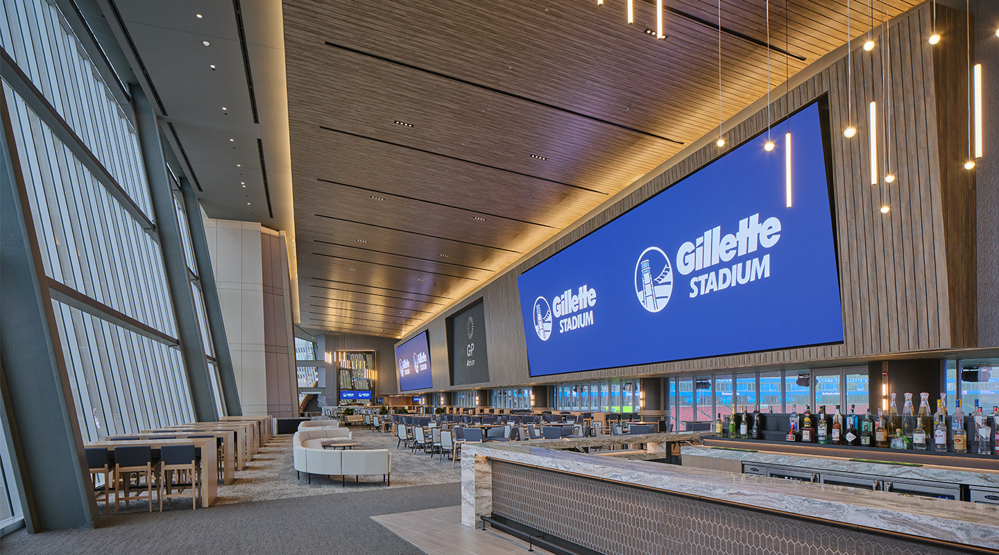 A modern lounge with high ceilings, large digital screens displaying Gillette Stadium, rows of seating, tables, and a bar with various bottles, all illuminated by natural light from tall windows on the left.