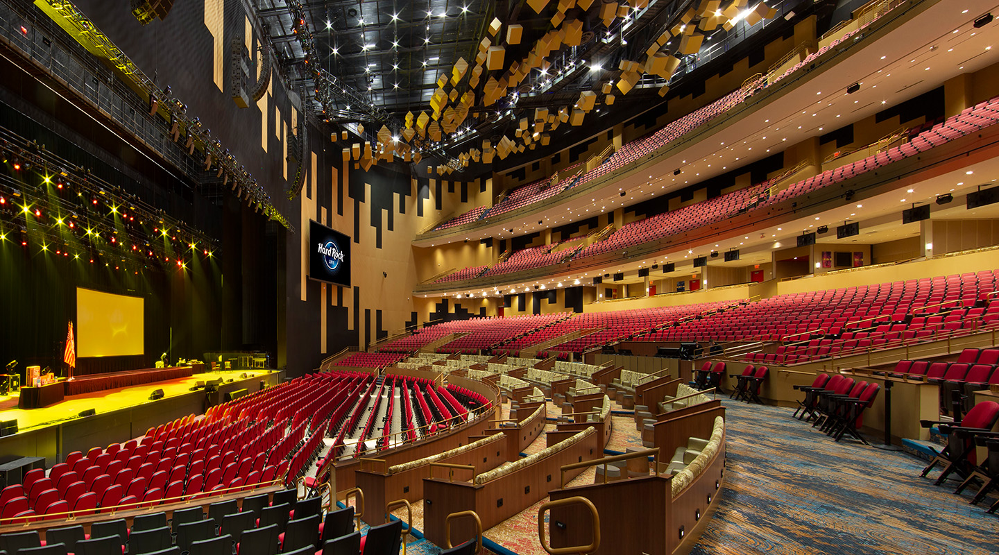 A spacious, modern theater with rows of red seats, curved balconies, and a large stage lit by yellow spotlights. The ceiling features geometric acoustic panels, and the walls are designed for optimal sound experience.