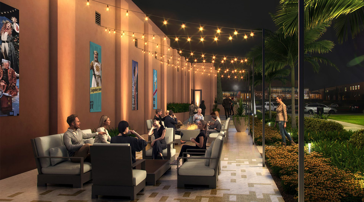 A group of people sit and converse on outdoor sofas under string lights at night; colorful posters hang on a wall behind them, and lush greenery borders the patio area.