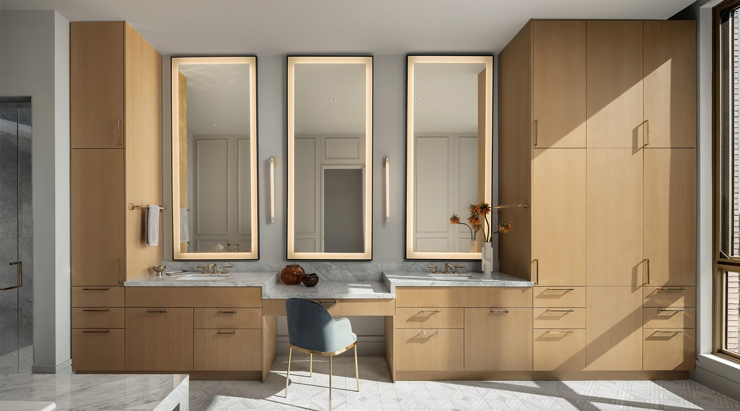 Modern bathroom with light wood cabinetry, three tall illuminated mirrors, two sinks with marble countertops, a central vanity area with a blue chair, and decorative vases, illuminated by natural light from a large window.