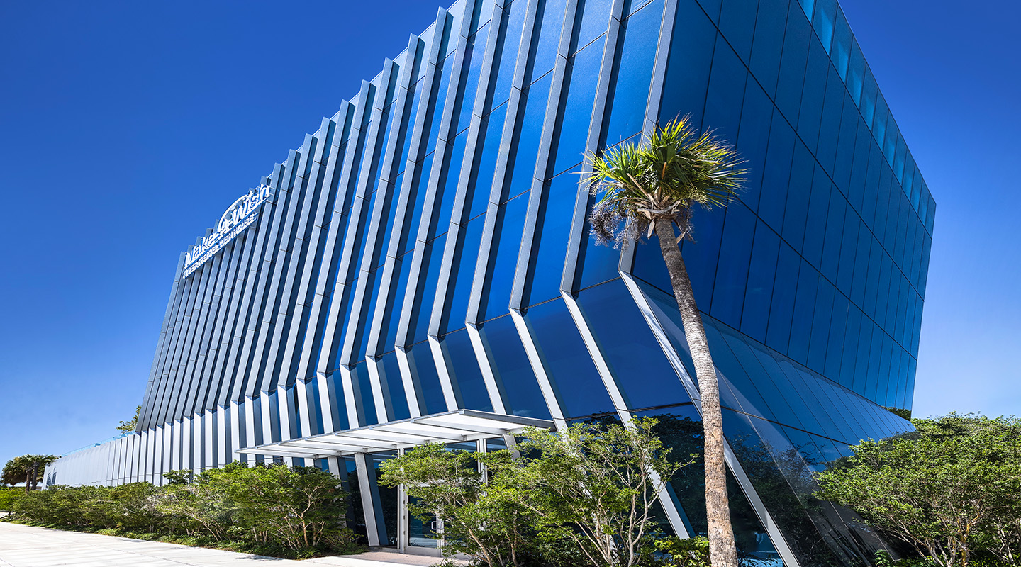 A modern, angled blue glass building stands against a clear sky, with a tall palm tree and green shrubs near the entrance. The building reflects the blue of the sky, creating a sleek, contemporary appearance.