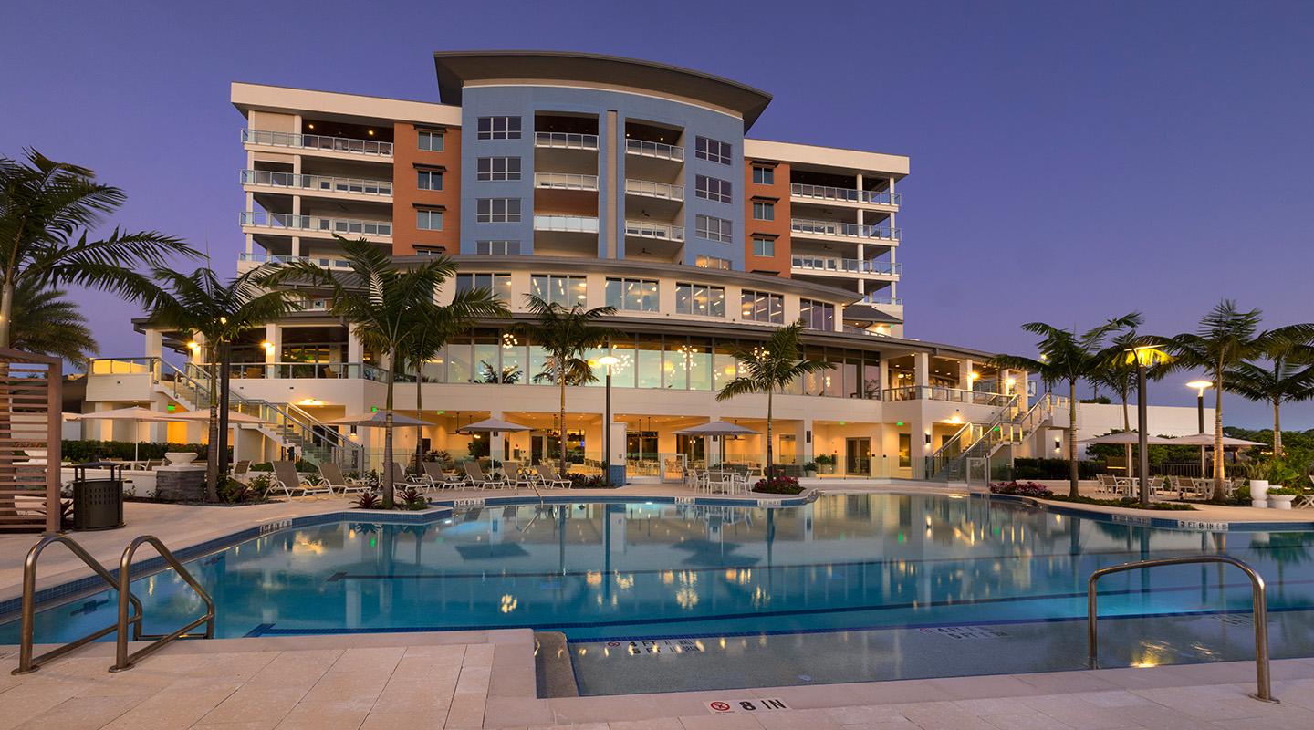 A modern, multi-story resort building with balconies is lit up at dusk, reflected in a large outdoor swimming pool surrounded by palm trees, lounge chairs, and patio tables.