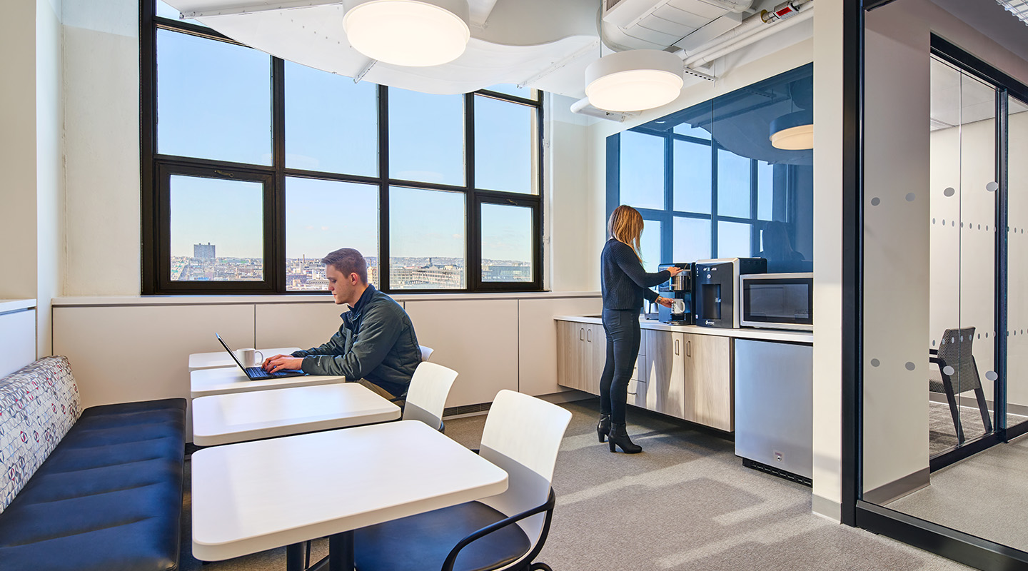 A man works on a laptop at a table in a modern office kitchen, while a woman prepares a drink at the counter near a microwave. Large windows show a city view in the background.