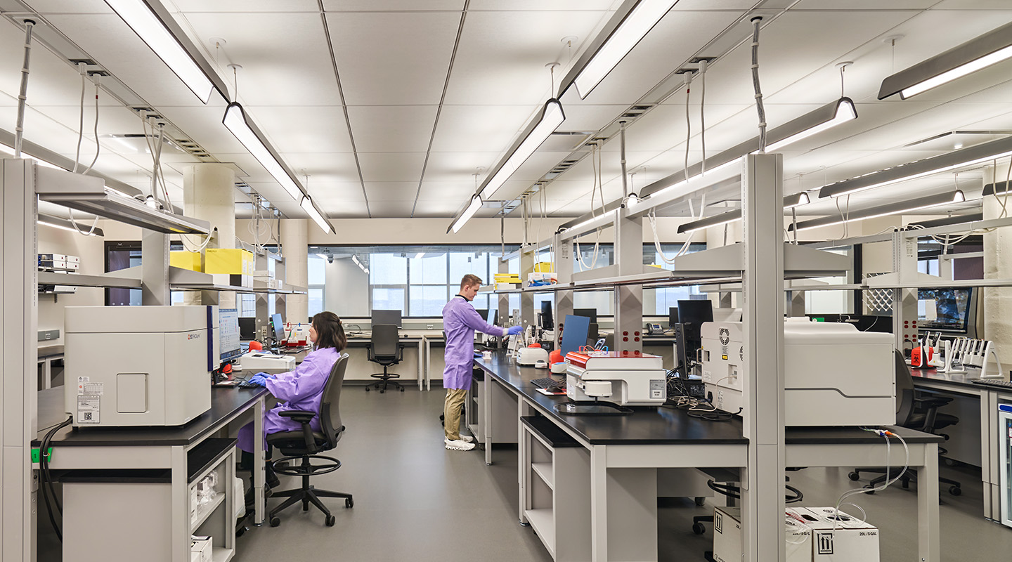 A modern laboratory with two people in purple lab coats working at separate stations with scientific equipment under bright overhead lights.