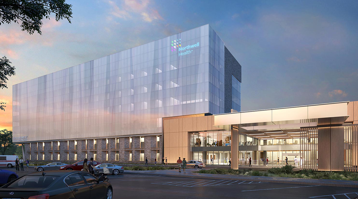 Modern hospital building with glass facade at sunset, displaying the Northwell Health logo on the upper corner. Cars are parked in front, and the entrance is well-lit, creating a welcoming atmosphere.
