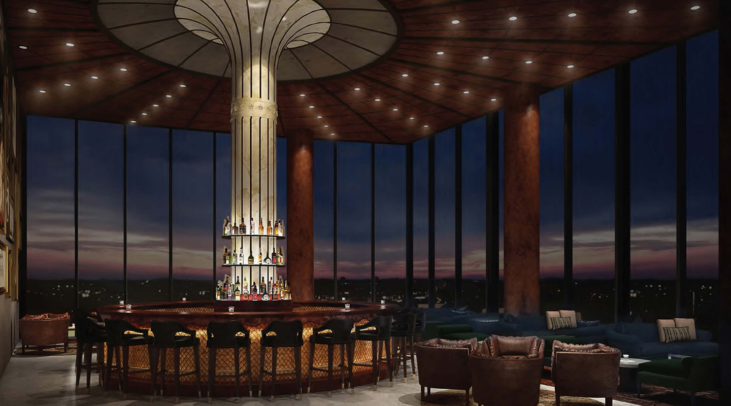 A stylish, modern bar with a circular counter surrounding a tall, illuminated column, featuring shelves of liquor. Large floor-to-ceiling windows reveal a nighttime cityscape and dark blue sky. Plush chairs and sofas are arranged nearby.