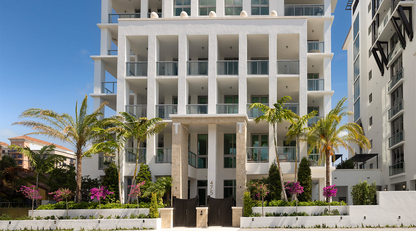 A modern white apartment building with balconies and large columns, surrounded by palm trees and vibrant landscaping, under a clear blue sky. The entrance features the number 475.