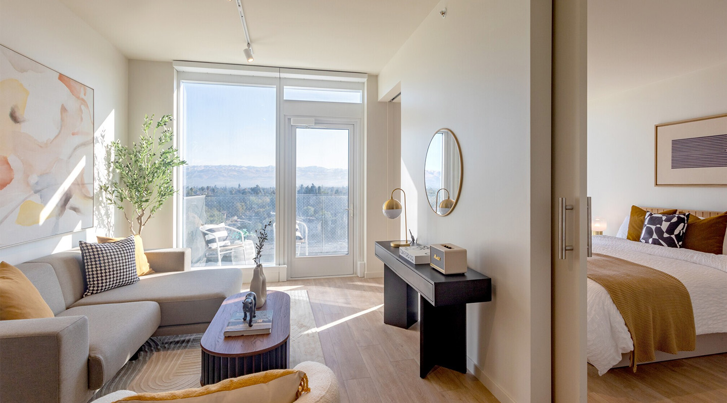 Modern, sunlit studio apartment with a gray sofa, abstract art, round coffee table, and a desk. Large windows open to a balcony with mountain views. A cozy bed with gold accents is visible in a corner.