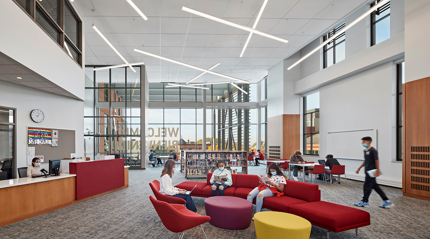 A modern library with high ceilings, large windows, and bright lighting. Several people, some wearing masks, are reading or working at tables, while others sit and talk on red chairs and couches in a lounge area.