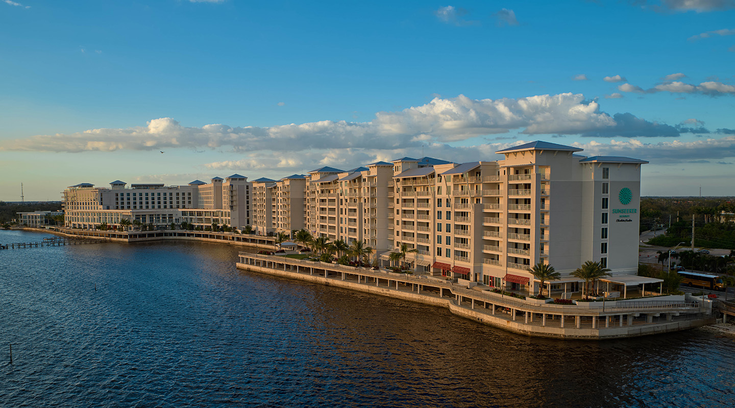 A large waterfront hotel complex with multiple connected white buildings featuring balconies, surrounded by palm trees, sits beside calm water under a blue sky with scattered clouds at sunset.