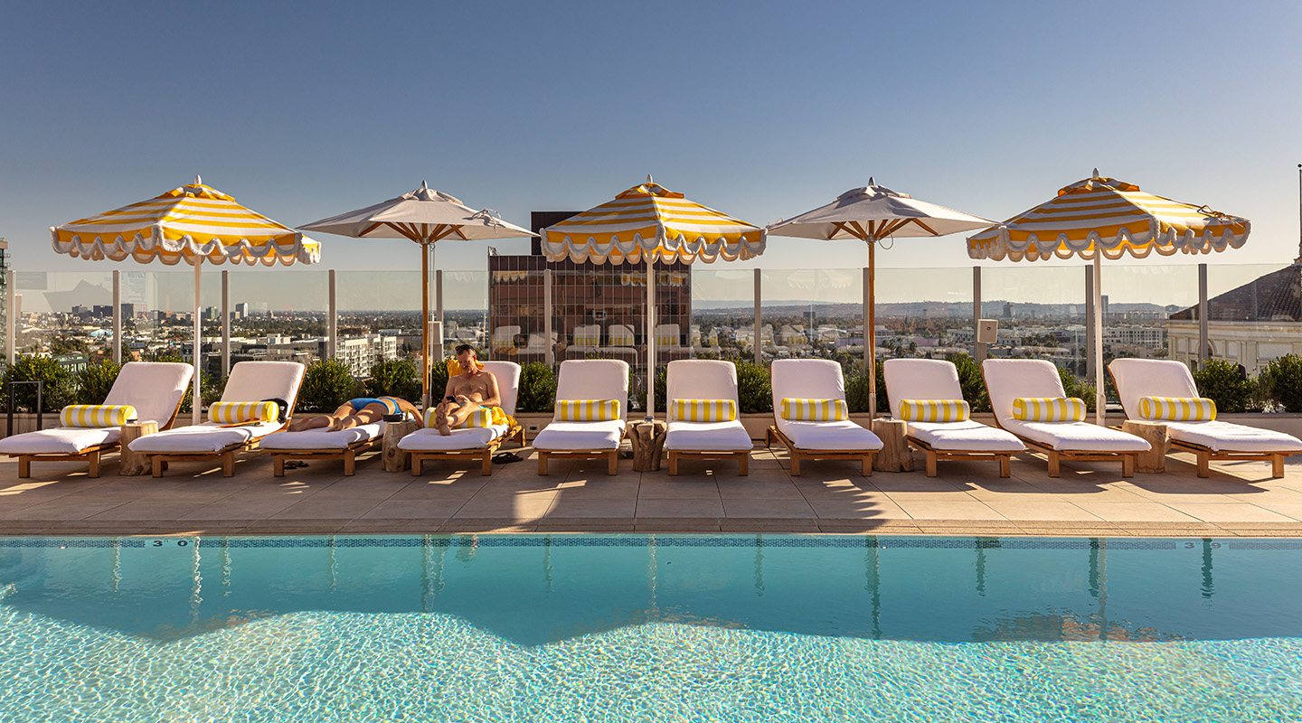 A rooftop pool with clear blue water, lined with white cushioned lounge chairs and yellow striped umbrellas. A person relaxes on one chair, with a cityscape visible in the background under a clear sky.