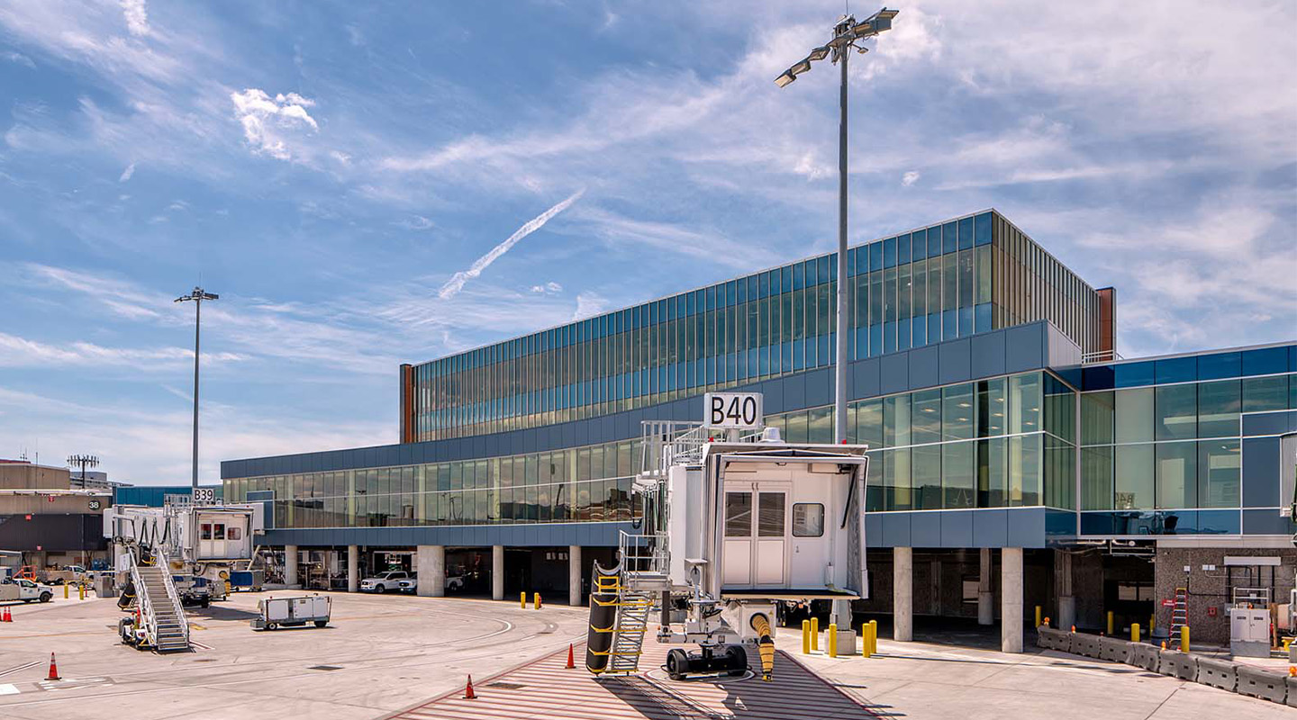 A modern airport terminal with large glass windows and gate B40 visible. Jet bridges and service vehicles are on the tarmac under a blue sky with scattered clouds.