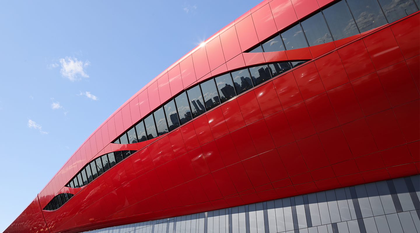A modern, futuristic building with bold, smooth red panels and curved, wave-like windows under a clear blue sky. The sleek architecture reflects sunlight, creating a dynamic and vibrant look.