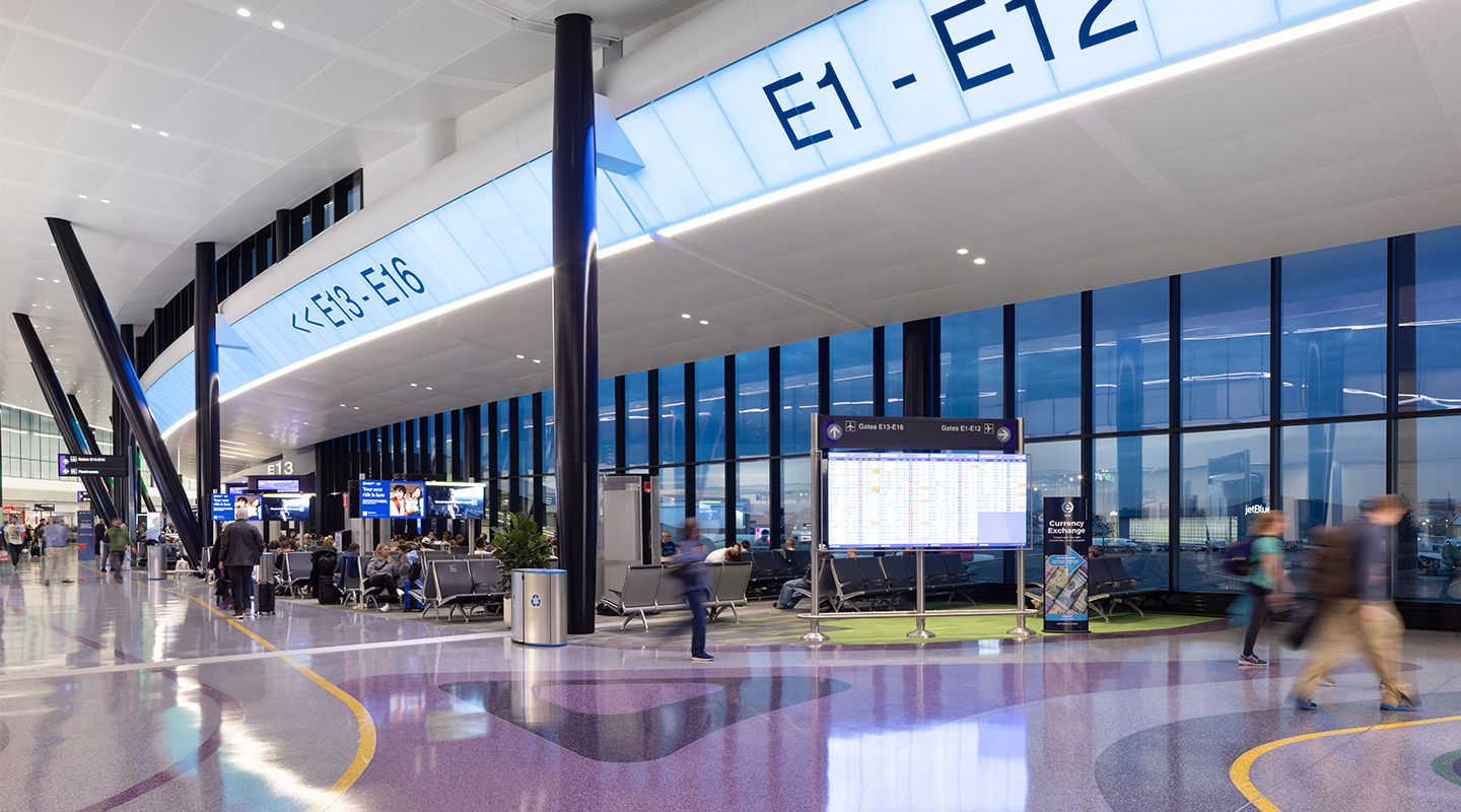 A modern airport terminal with people walking and waiting near gates E1-E16. Large windows, bright lighting, digital screens, and a glossy floor are visible, creating a spacious and contemporary atmosphere.