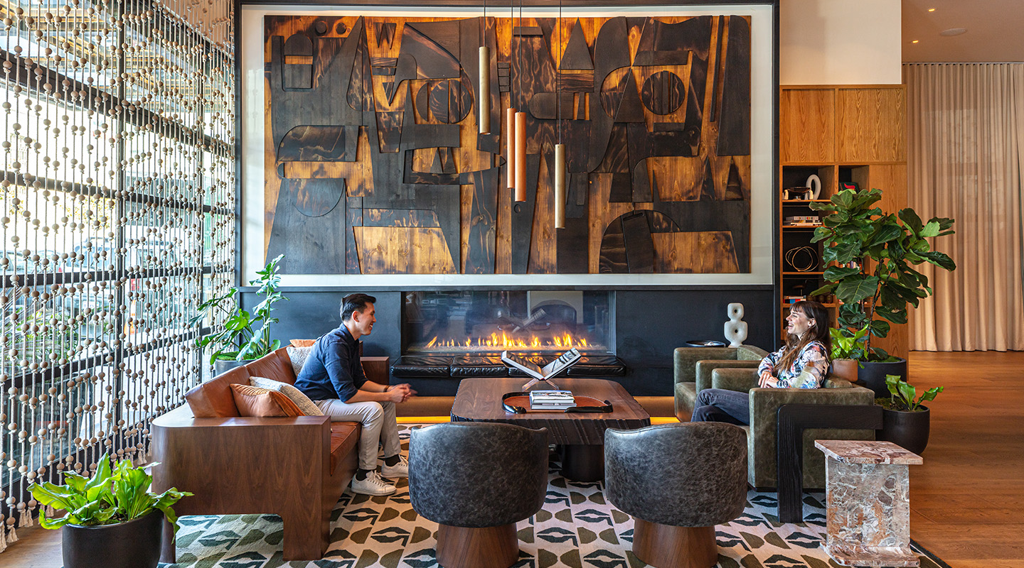 Two people sit and converse in a stylish lounge with modern furniture, large abstract art above a fireplace, indoor plants, and warm natural light streaming through tall windows.