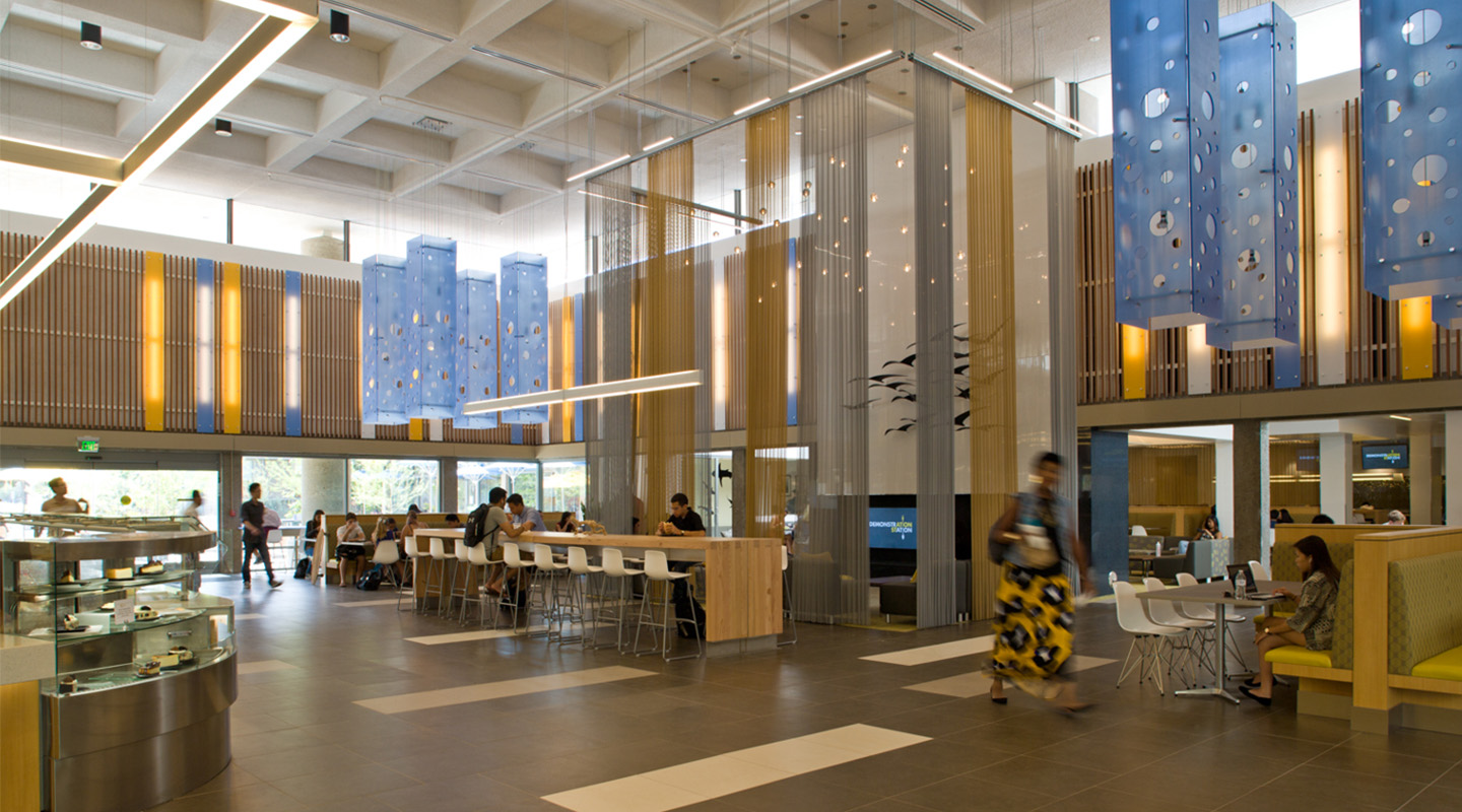 Modern, spacious cafeteria with high ceilings, large windows, blue hanging decorations, and groups of people sitting at tables and counters. A glass display case with food is visible on the left.