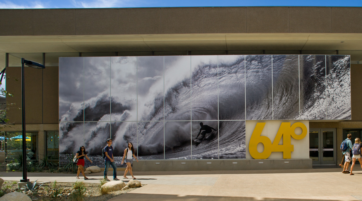 A large black-and-white mural of a surfer riding a wave covers a building’s exterior. Four people walk past in front, and large yellow numbers “64°” are displayed at the building’s entrance.