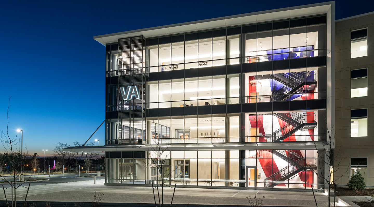 A modern, glass-front VA building at dusk, with an illuminated staircase showing a large American flag design inside. The building is well-lit, and the sky is deep blue.