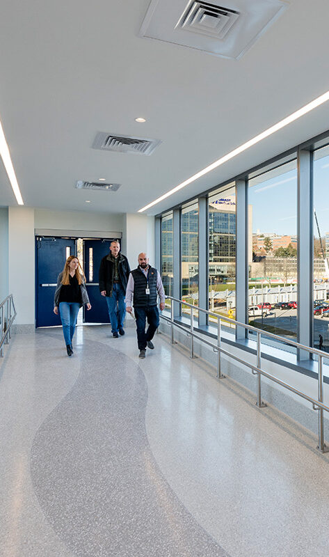 A modern enclosed skywalk with large windows shows several people walking inside, with views of a cityscape, office buildings, and a parking lot visible outside on a clear, sunny day.