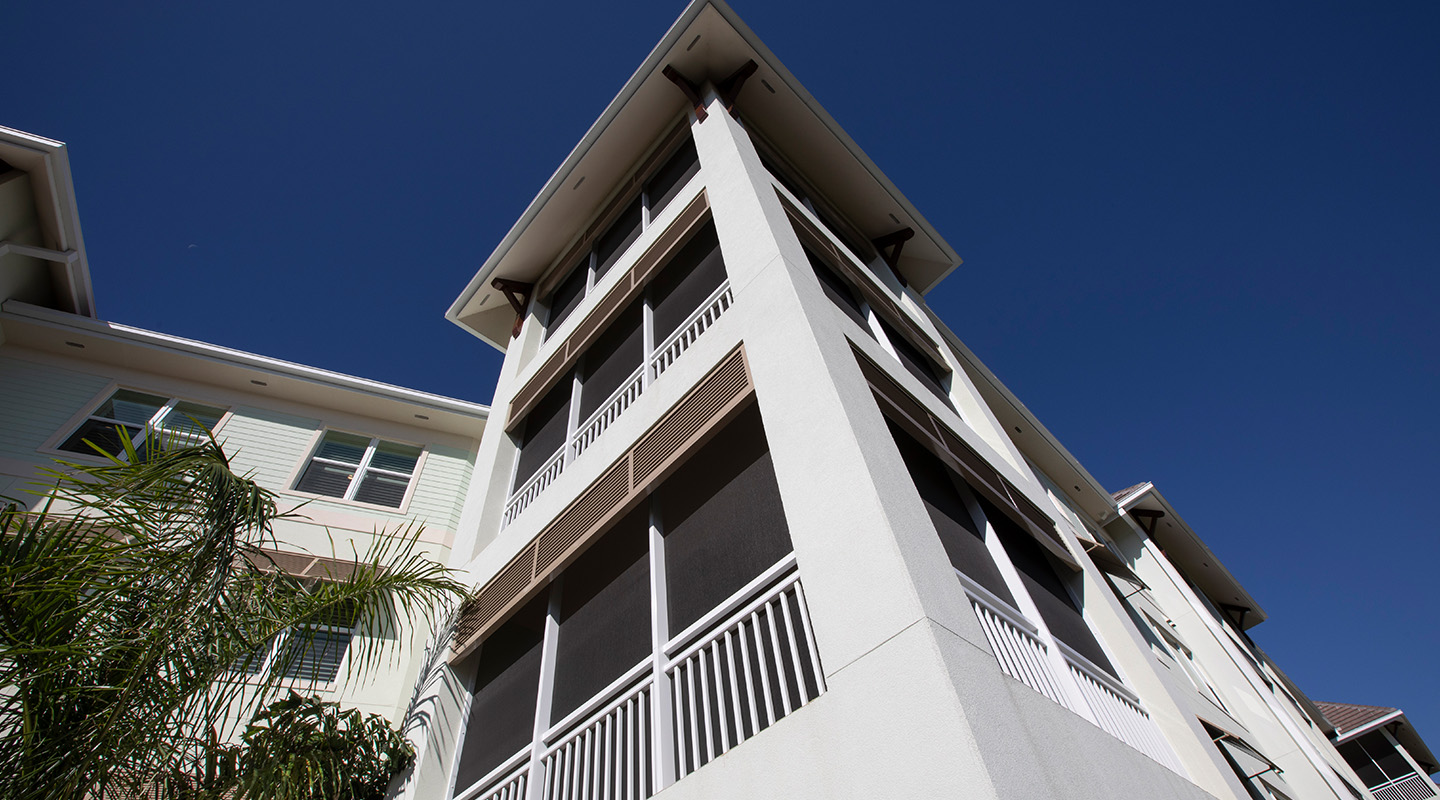 A low-angle view of a modern apartment building with white walls, screened balconies, and a clear blue sky in the background. A palm tree is visible to the left.