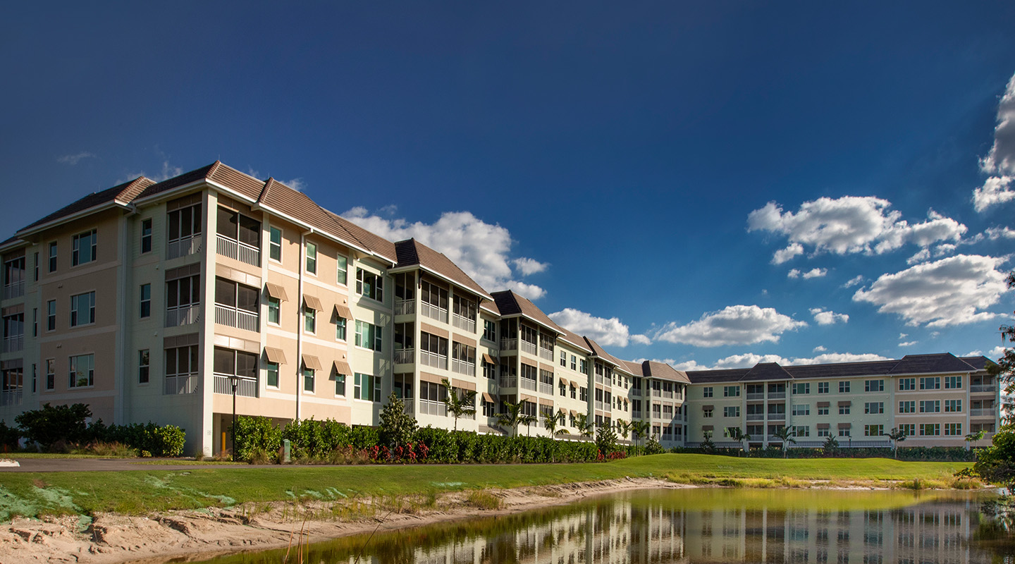 A modern three-story apartment building with light-colored walls and balconies, surrounded by greenery, under a blue sky with clouds, and reflected in a small pond in the foreground.