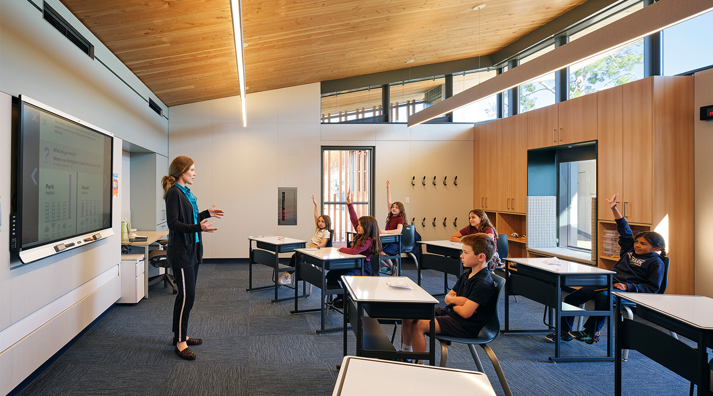 A teacher stands at the front of a modern classroom, speaking to students seated at individual desks. Several students have their hands raised, and large windows allow natural light to fill the room.
