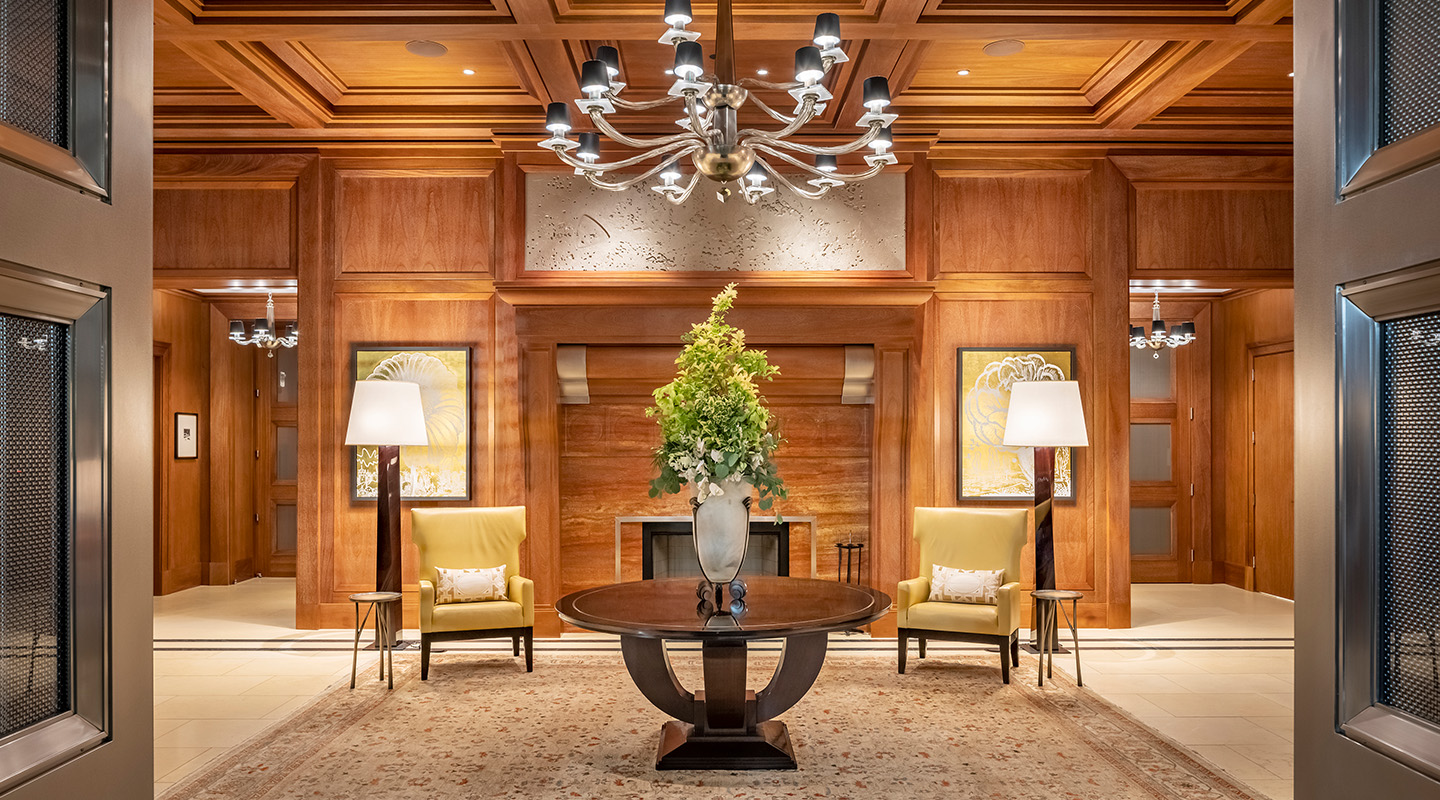 A luxurious hotel lobby with wood-paneled walls, a chandelier, two yellow armchairs with floor lamps, artwork on the walls, and a round table with a large plant centerpiece on a patterned rug.
