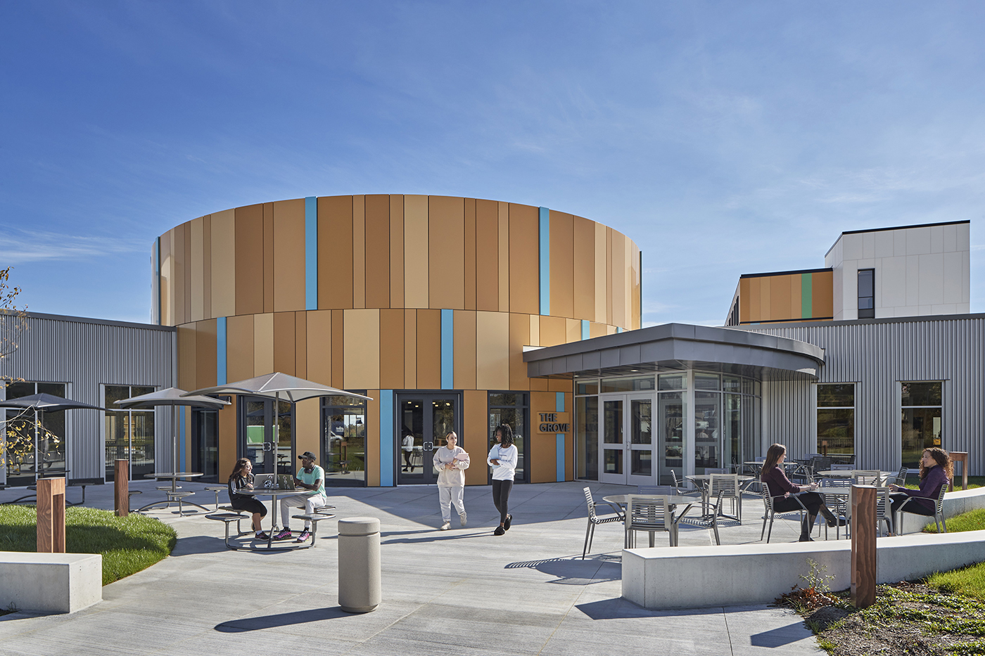 People sit and walk outside a modern building with round and angular sections, featuring tan, blue, and gray panels. Several tables with umbrellas are on a sunny patio, and the sky is clear and blue.