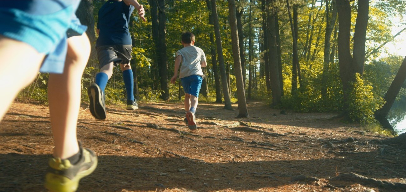 Three children are running along a sunlit forest trail, surrounded by tall trees and greenery. The scene is lively and energetic, with sunlight filtering through the leaves onto the path.