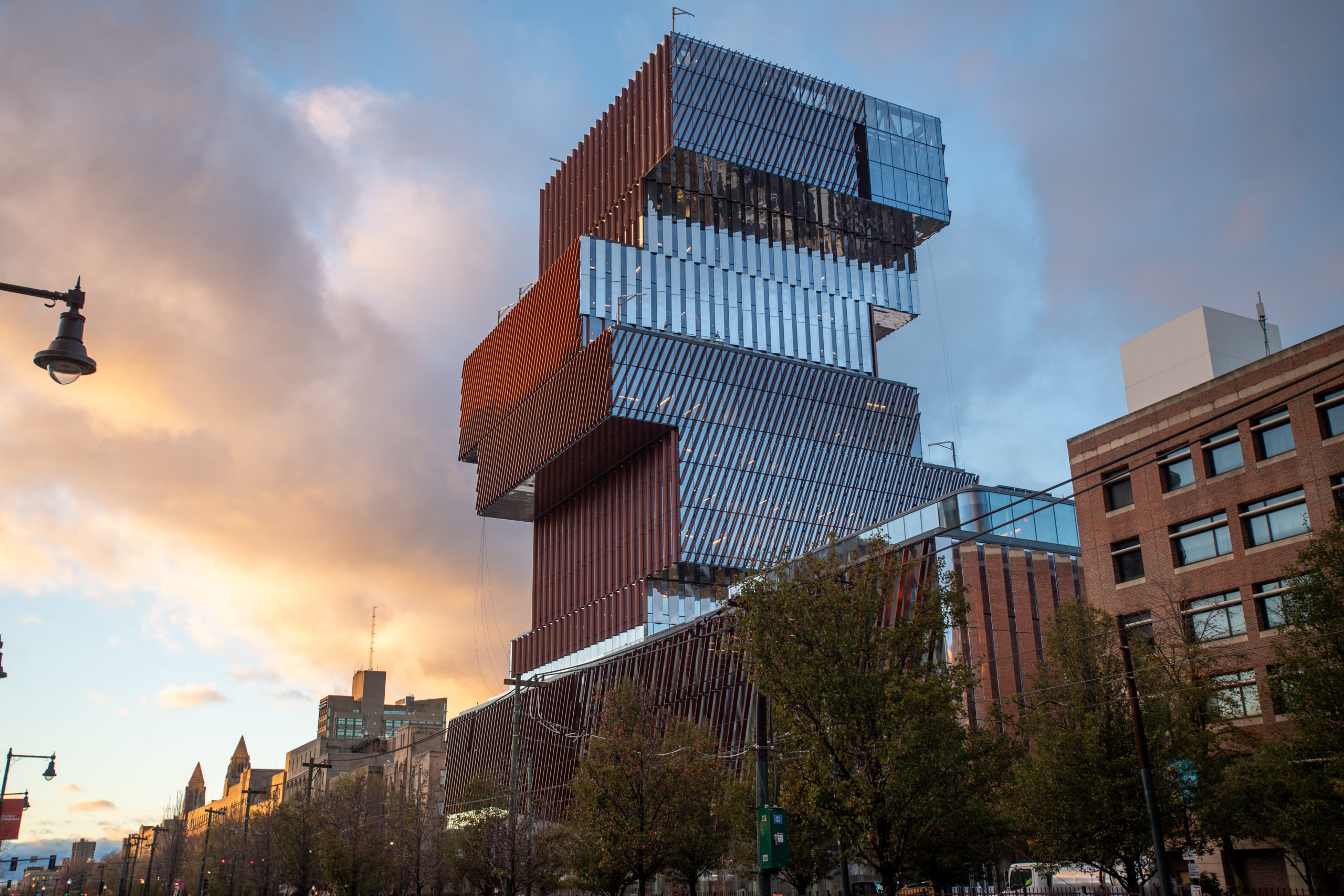 A modern, stacked glass and metal building with angular, offset sections rises above a city street at sunset, surrounded by trees and older brick buildings.