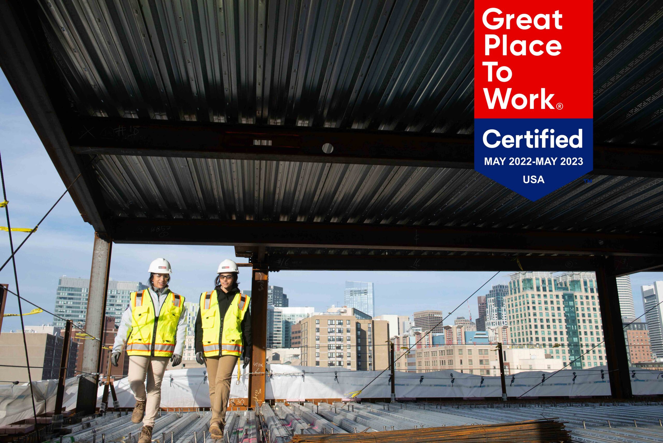 Two construction workers in safety gear walk at a building site with a city skyline in the background. A Great Place to Work Certified May 2022-May 2023 USA badge is displayed in the upper right corner.