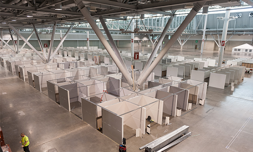 Large indoor convention space filled with rows of temporary partitioned cubicles, likely for use as makeshift medical stations or shelters. Few workers and construction materials are visible in the foreground.