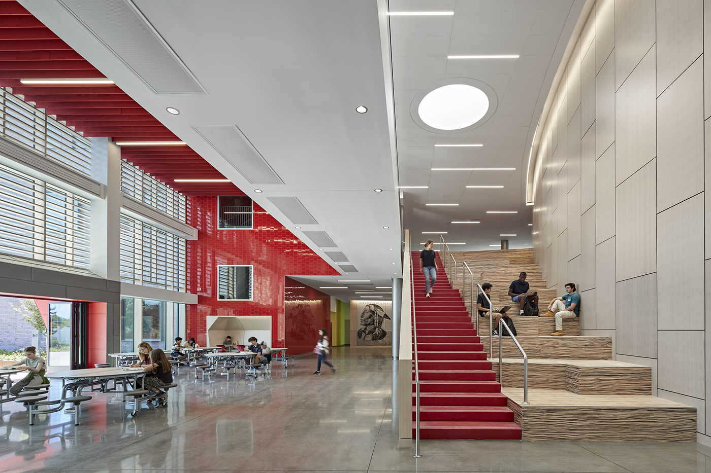 Modern, spacious indoor area with high ceilings, red accents, large windows, and a wide red-carpeted staircase; people are sitting and walking, and others are seated at tables, studying or socializing.