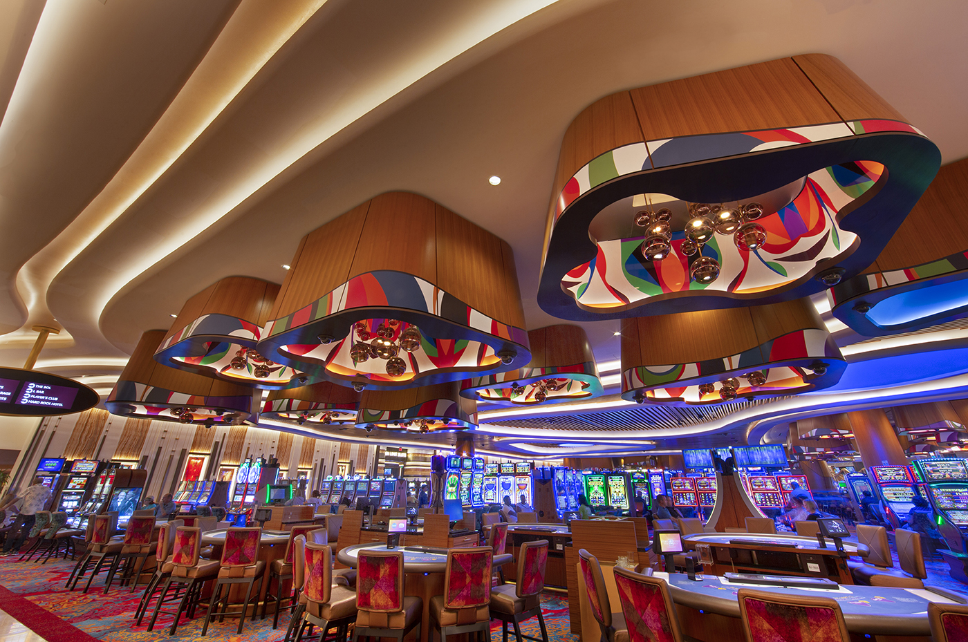 A vibrant casino interior featuring colorful, artistic ceiling lights, rows of slot machines, and red-upholstered chairs around gaming tables under warm lighting.