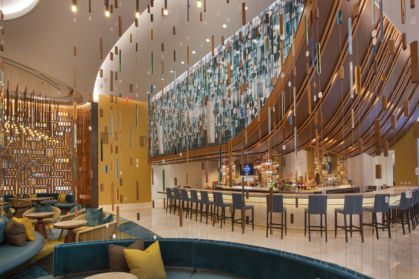 A modern hotel bar with stylish hanging light installations, a long curved bar with high chairs, blue and gold seating, and decorative wall panels, creating a vibrant, luxurious atmosphere.