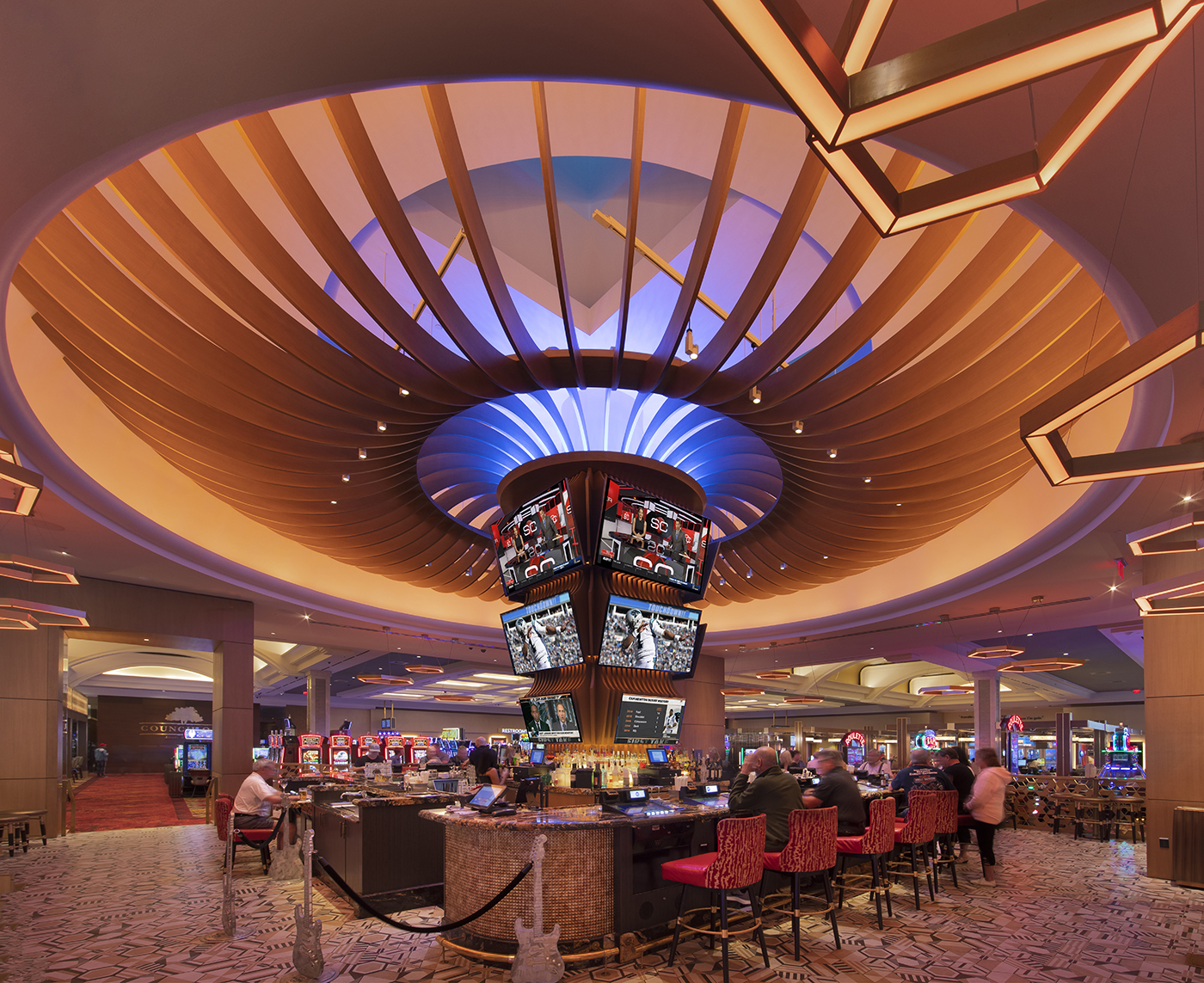 A round bar with red chairs and multiple TVs sits under a large, modern ceiling with dramatic lighting in a lively casino filled with slot machines and gaming tables.