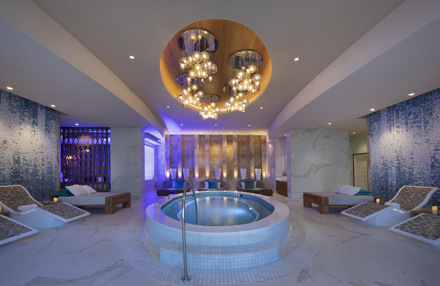 A luxurious indoor spa with a central round hot tub, lounge chairs, candles, ambient blue lighting, textured blue and white walls, and modern chandeliers hanging from a wooden ceiling feature.